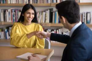 Happy Indian student woman shaking hands with male teacher in library, thanking for tutorial, training. Positive young female employee giving handshake to boss at meeting, smiling, laughing